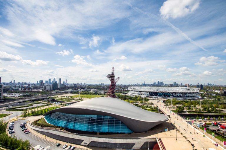 ChatGPT sa: West Ham Uniteds London Stadium og London Aquatics Centre i forgrunnen, med Canary Wharf og City of London på horisonten, sett fra Stratford.
