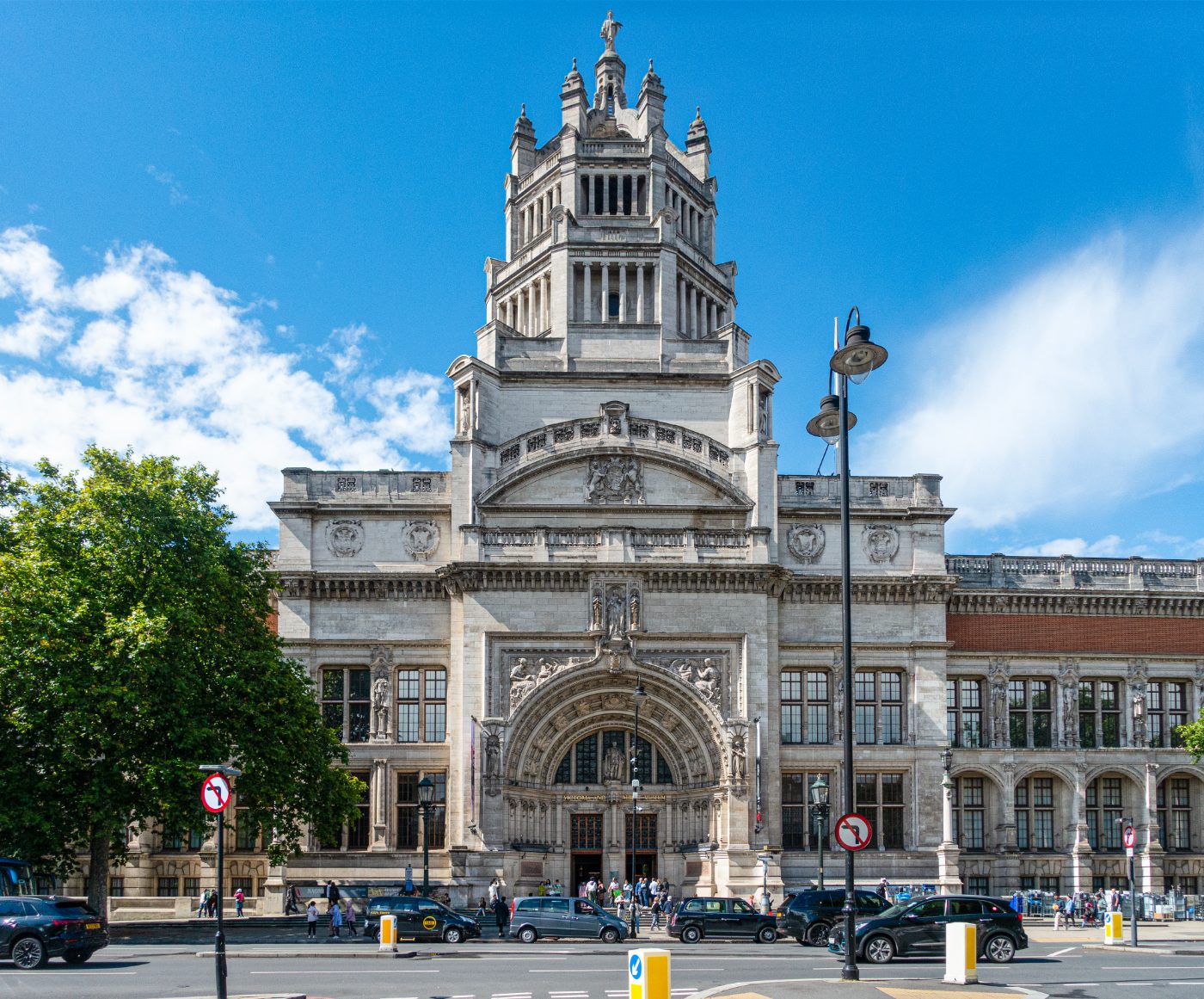 Hovedinngangen til Victoria and Albert Museum (V&A) i South Kensington, London, med fotgjengere, besøkende og drosjer utenfor. Fasaden, designet av Aston Webb i Portland-stein, har statuer av dronning Victoria, prins Albert og Fame på tårnets krone.
