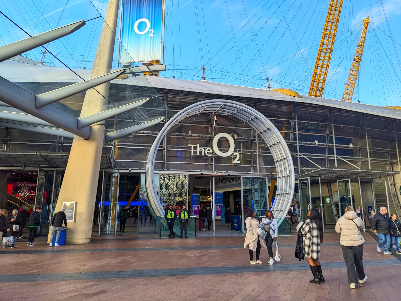 Besøkende ved hovedinngangen til The O2 Arena under de karakteristiske gule mastene på en solrik dag.