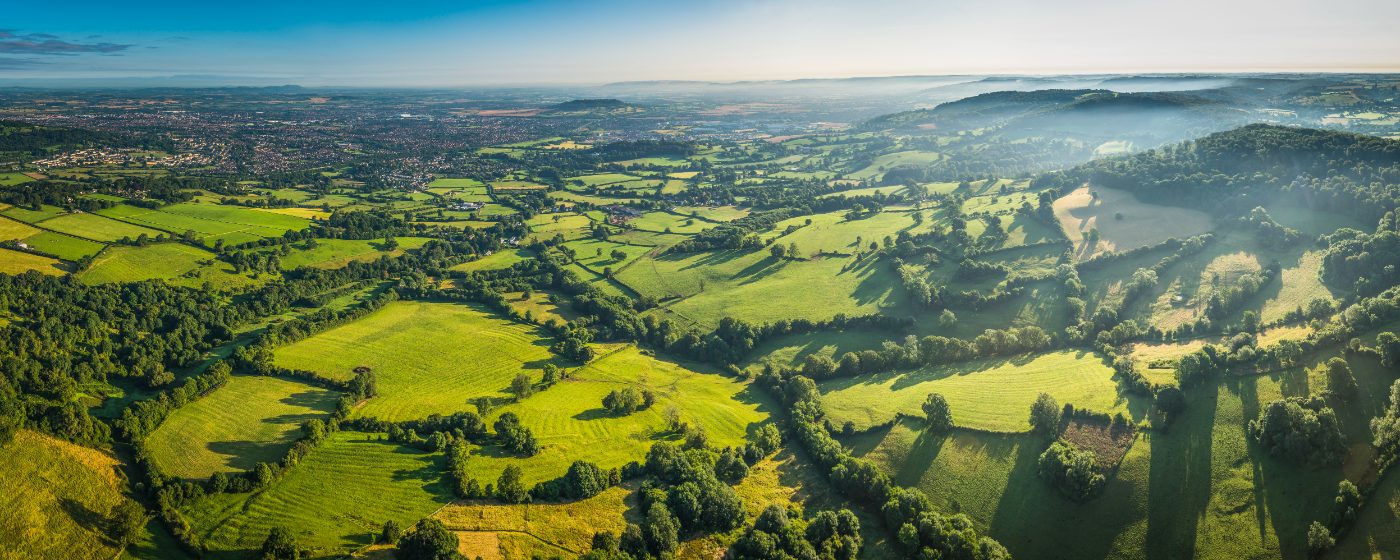 Panoramisk flyfoto over Cotswolds med grønne jorder, tåkete åser og en landsby.