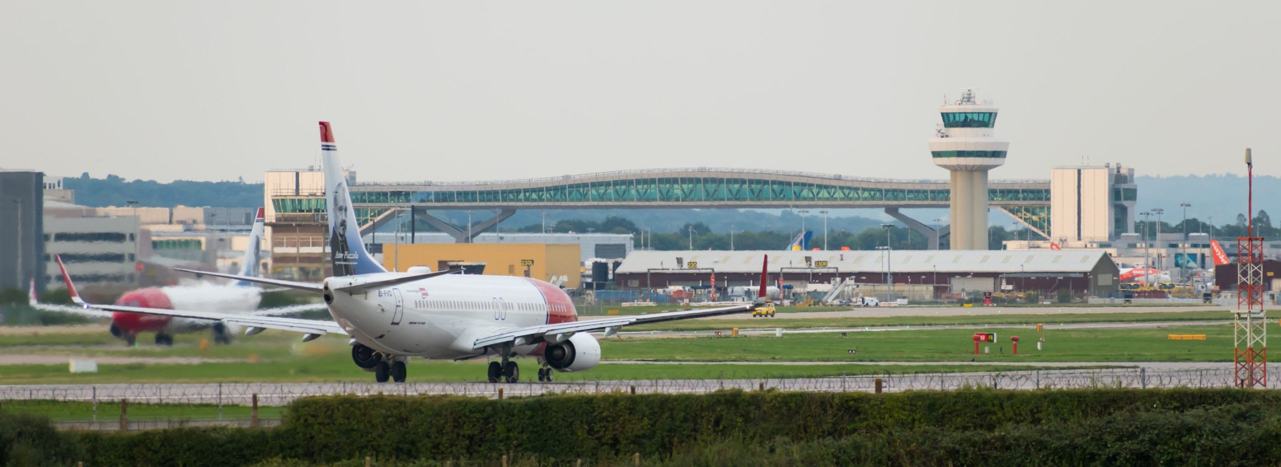 Norwegian Airlines-fly gjør seg klare til avgang fra Gatwick lufthavn, med flyplassens kontrolltårn og flybro i bakgrunnen.